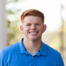 A man standing outside with red hair and a blue shirt infront of a pole
