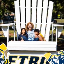 A mom and two children sitting in a giant adirondack chair on a sunny day.