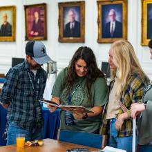 Four people looking at a yearbook in a room with portraits in gold frames on the wall. 