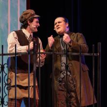 Two male actors singing on stage behind an iron fence. 