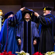 Three men in graduation regalia with the left and right men hooding a doctoral student in the middle.