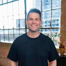 Man standing in front of windows in a black t-shirt and khaki pants.