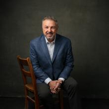 A man in formal clothing sitting in a wooden chair in front of a grey backgrop