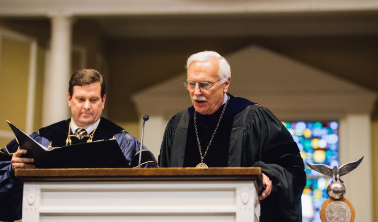 President Dr. Blackburn speaking at poduim with others dressed in academic regalia during graduation ceremony. 