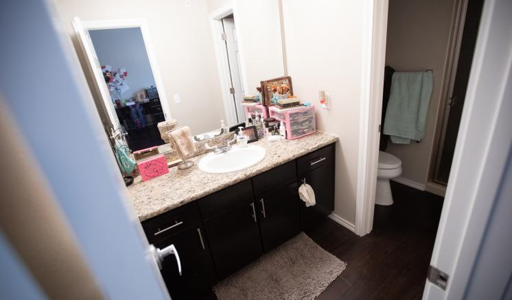 A bathroom with a sink, mirror, and a countertop with toiletries and cosmetics. Dark brown cabinets are below the counter, and a toilet is seen in a room to the right.