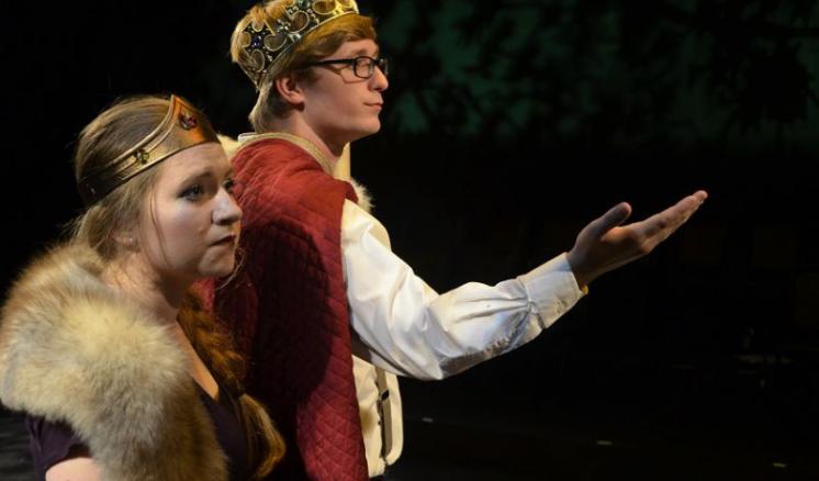 Two actors in royal costumes, one with a crown and fur, the other with a crown and cape, on stage.