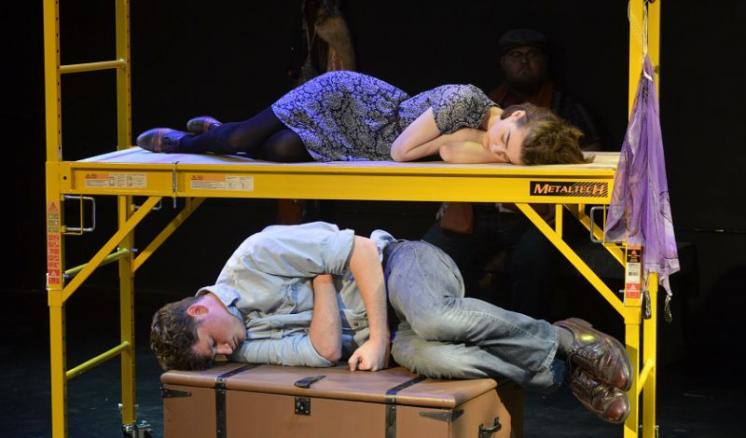 Two people sleeping on a yellow scaffolding platform and a brown box in a dark theater scene.