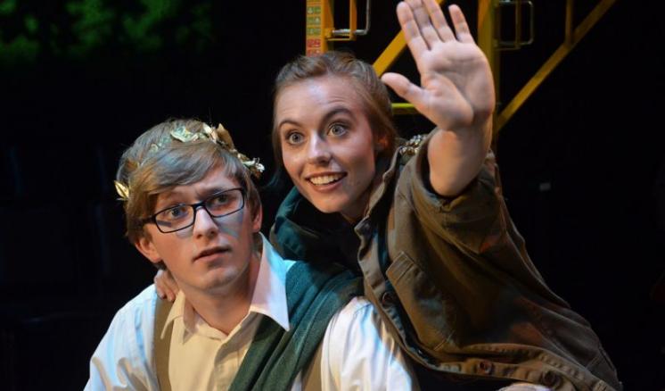 Two people on stage, one with glasses and a gold headpiece, the other raising their hand.