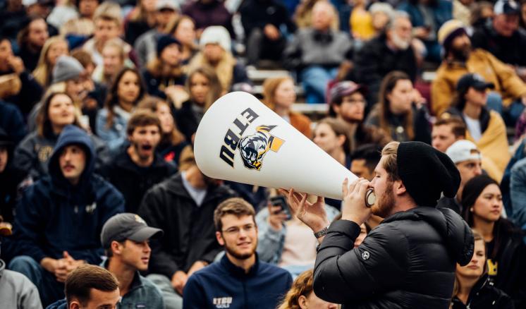 A man uses a large megaphone to address a crowd at an outdoor event. The megaphone displays an "ETBU" graphic with a Tiger image.