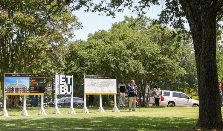 People in a park area with large signs and a white vehicle under trees.