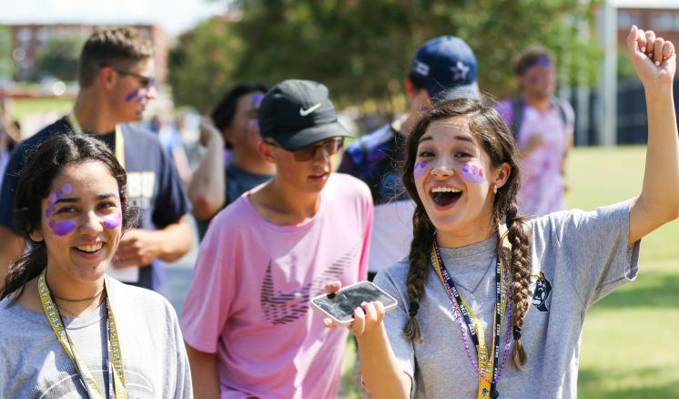 A group of young people smiling outdoors, with one holding a phone and raising their fist.