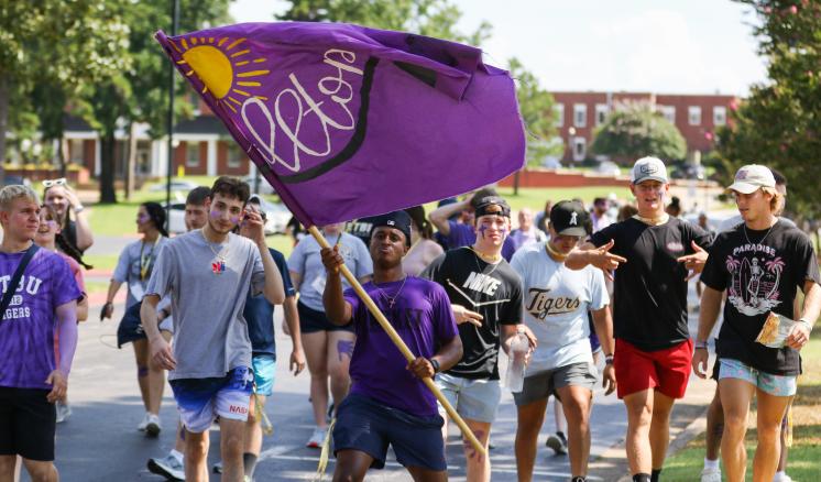 A group of people walking outdoors, with one person holding a large purple flag that has a yellow sun and the word "Rebels."