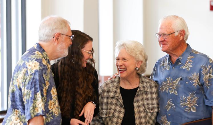 Four people laughing together indoors, two men in floral shirts and two women, one with a checkered shirt.