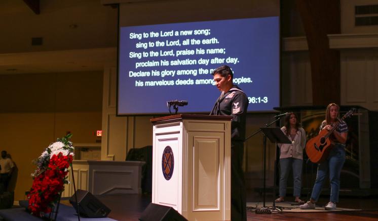 Person at a podium in a church with a background screen displaying text and musicians on stage.