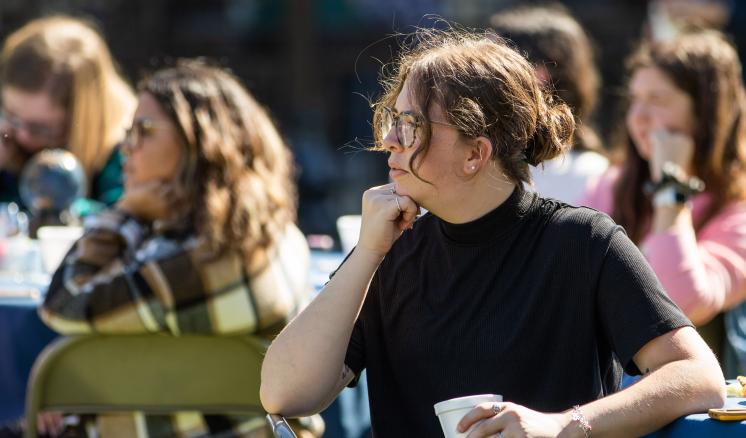 People seated outdoors, engaging in a casual event, with a person in front holding a paper cup.