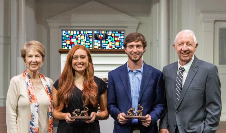 Four people standing indoors, two holding awards, with a stained glass window behind them.