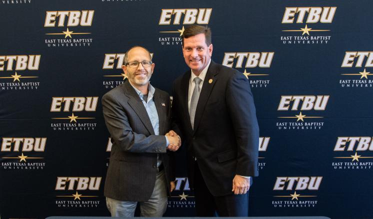 Two men shaking hands in front of an "ETBU" backdrop with documents on the table.