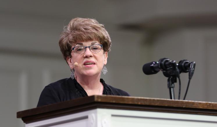 A person with short hair and glasses speaks at a podium with multiple microphones.
