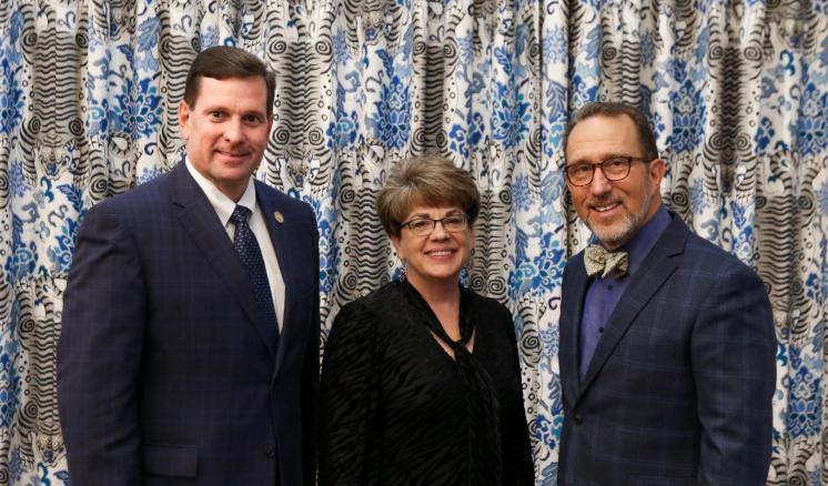 Three people standing in front of a patterned curtain, dressed formally.