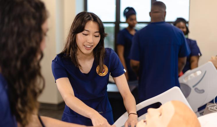 Person in a blue uniform practicing on a medical mannequin in a classroom setting with others in the background.
