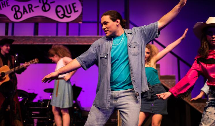 Performers dancing on stage with a "Bar-B-Que" sign above, set against a purple-lit background.