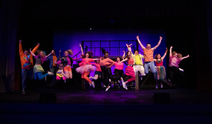 Performers in colorful outfits jumping on a stage with a vibrant purple and blue background.