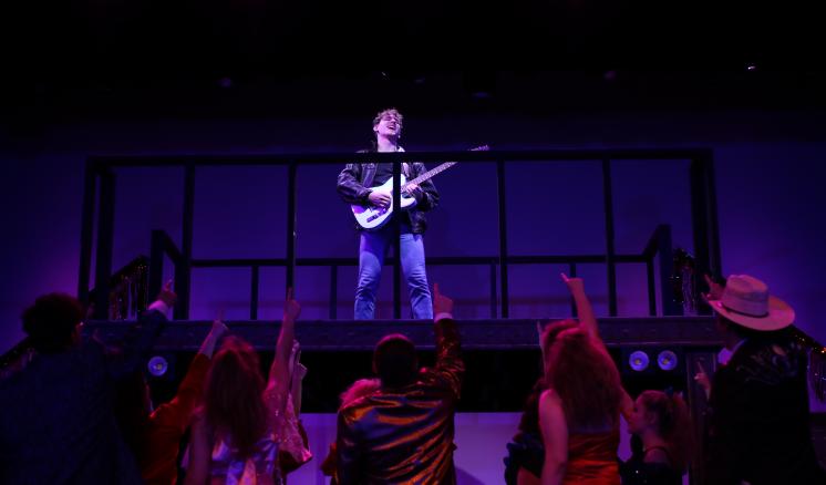 Stage performance with a guitarist on a platform under pink and purple lights, with a cheering audience below.