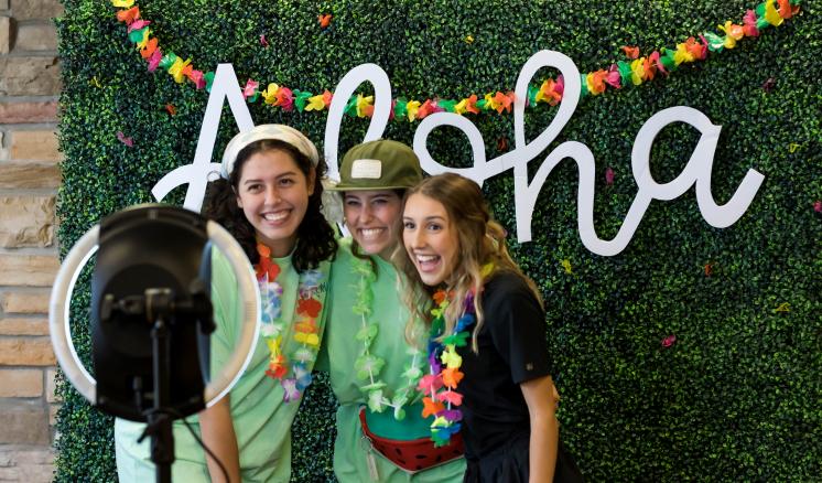 2022_connexus_img_1 Three women taking pictures in front of a ring light with a plant backdrop with the word aloha in the back