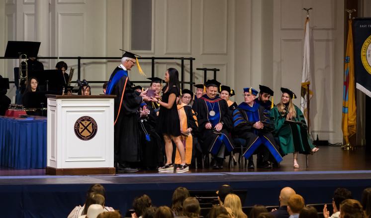 Man in graduation regalia giving a woman a plaque with others in the graduation regalia in the background
