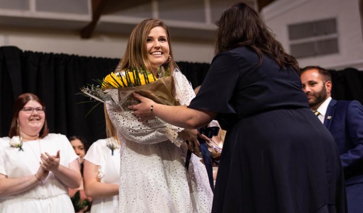 A woman in a white dress receives a bouquet of sunflowers from another woman in black, with people clapping in the background.