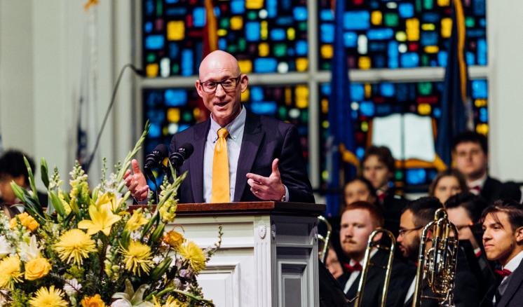 Man speaking behind a podium with people in the background and yellow flowers in the front