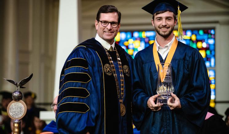 Two men smiling at the camera in graduation regalia with one holding an award