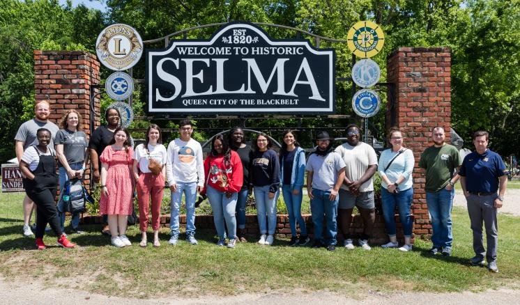 A group of nineteen people stands in front of a sign reading "Welcome to Historic Selma, Gateway to the Blackbelt," with trees in the background.