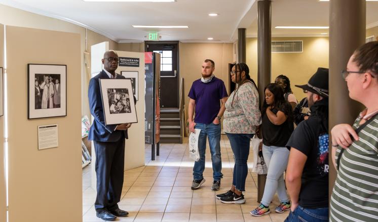 A man in a suit presents a framed photo to a group in a gallery space.