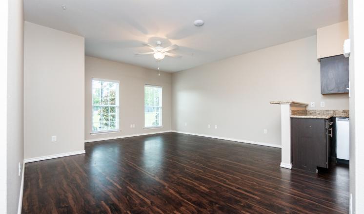 Empty room with dark hardwood flooring, light beige walls, two large windows, and a kitchenette area.