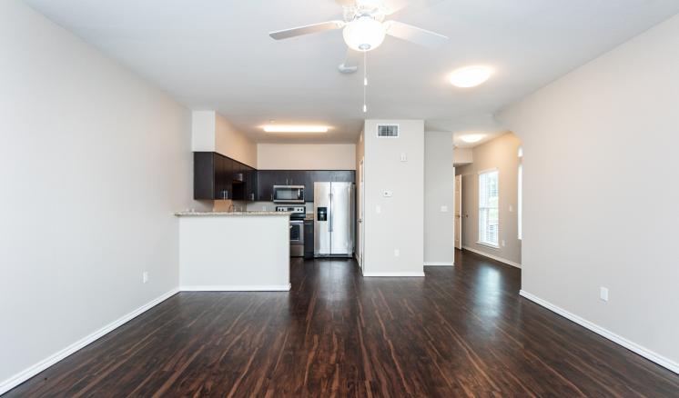 Empty apartment interior with dark wooden floors, open kitchen, and a ceiling fan.