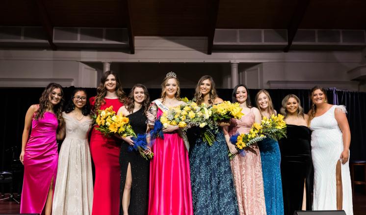 A group of women in ballgowns smiling at the camera on stage