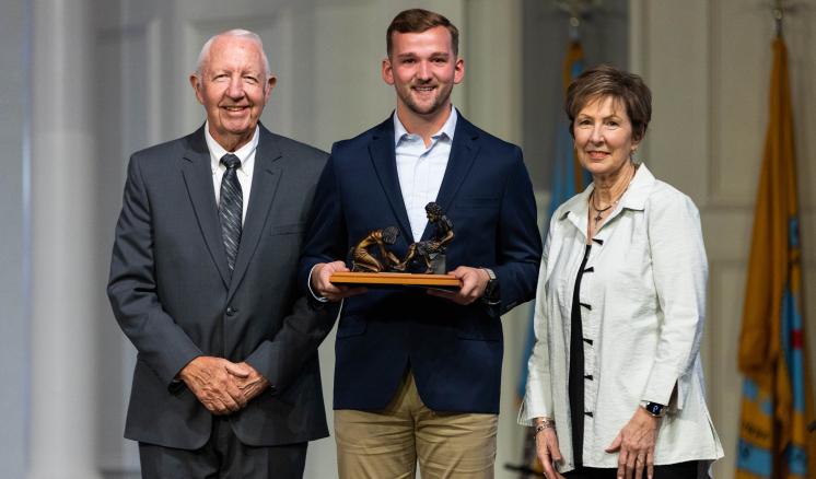 Two men and a women smiling at the camera in a chapel with the middle person holding an award