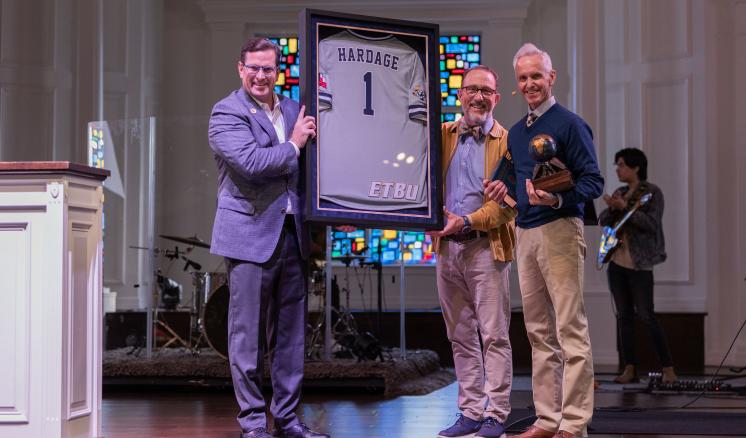 Three men smiling at the camera holding a jersey up with the number one on it on a stage