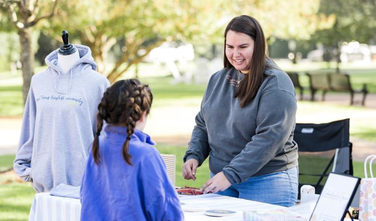 A woman smiling down at a little girl behind a table with a jacket on top of the table outside