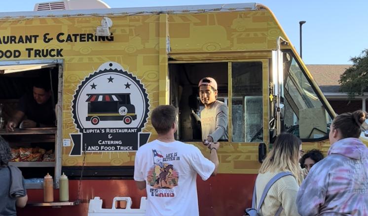 Man reaching into food truck window to give the man in the truck something with people standing on the right