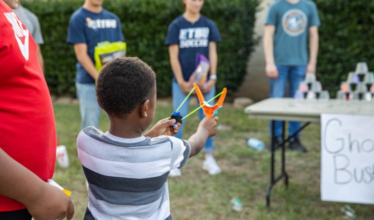 A little boy holding a sling shot trying to knock down cups on a table