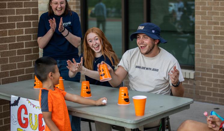2022_misd_fall_fest_0725 A man and woman sitting at a table with pumpkin cups smiling at a little boy with a woman clapping her hands in the background