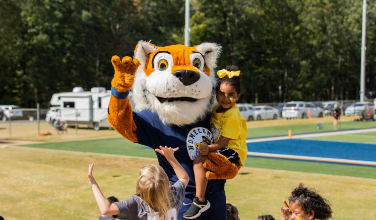 Tiger mascot holds a smiling girl in a yellow outfit, surrounded by excited children near a sports field.