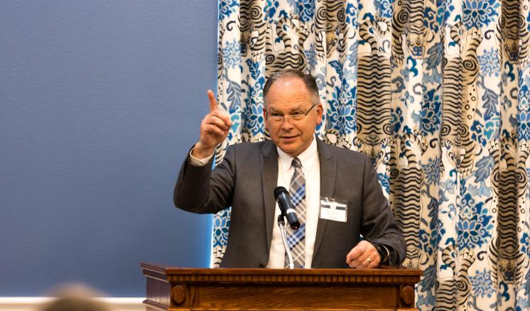 A man behind a podium looking out at the crowd with his hand out and his pointer finger pointing