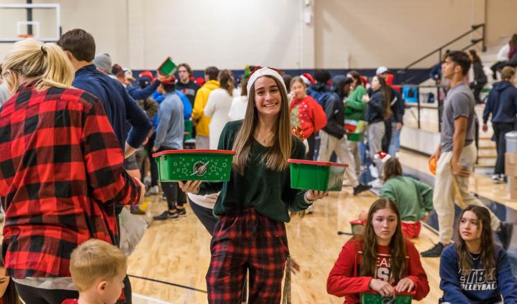 2022_operation_christmas_child_img_1 Woman smiling at camera with christmas boxes in hand with groups of people in the background