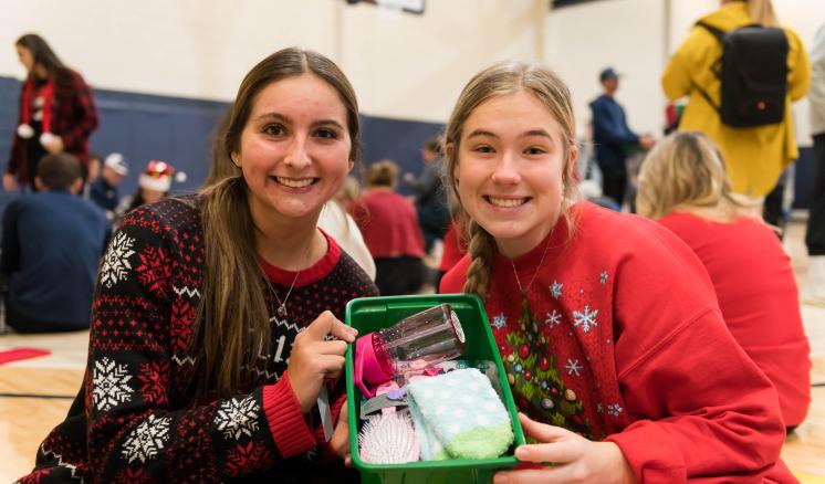 2022_operation_christmas_child_img_2 Two women smiling at the camera with a christmas box between them in a gym