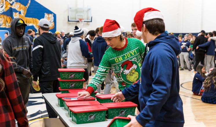 Two men in the foreground picking christmas boxes off a table with groups of people in the background in a gym