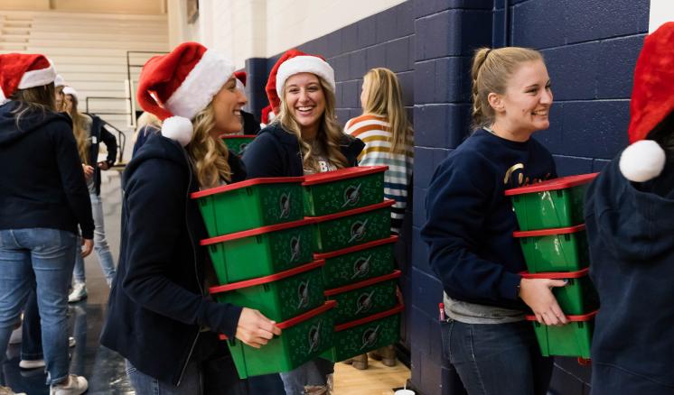 Three women walking with stacks of christmas boxes in a gym