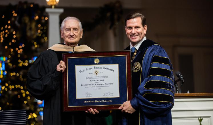 Two men in graduation regalia holding a framed diploma inside a chapel